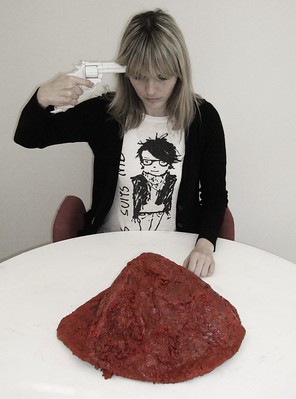 Woman with revolver aimed at her head in front of a giant plate of red goo.