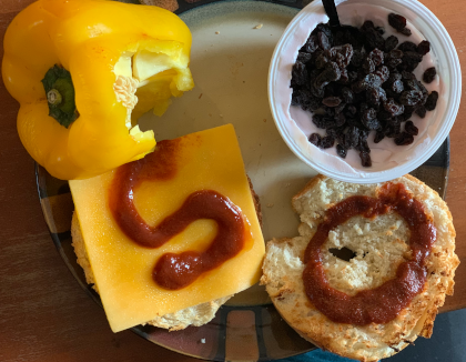 A breakfast consisting of: A partially eaten yellow pepper, yogurt with currents, and a bagel with cheddar cheese and a hot sauce 'S' on one side, and a hot sauce 'O' on the other.