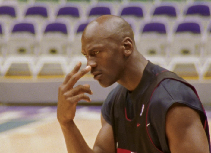 Michael Jordan sitting in an empty stadium pointing his index and middle finger at his eyes, staring at something with a determined look.