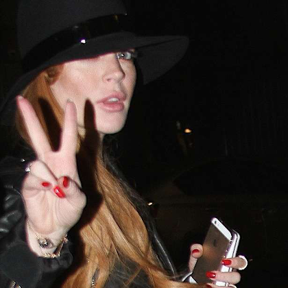 A redheaded woman in poor lighting holding a golden iphone 5 in one hand and showing the peace sign in the other. She's dressed in black with a black hat and painted red fingernails.