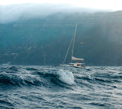 A sailboat with a wave taller than its mast towering, about to hit the boat.