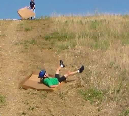 A man wearing a green shirt, black shorts, a blue bicycle helmet, and black hiking shoes with white ankle high socks sliding down a mostly dirt and weed hill, on his back, seemingly out of control.