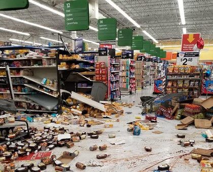 A picture of a Walmart store with fallen shelves, products scattered on the ground, and spilled liquids all over the floor.