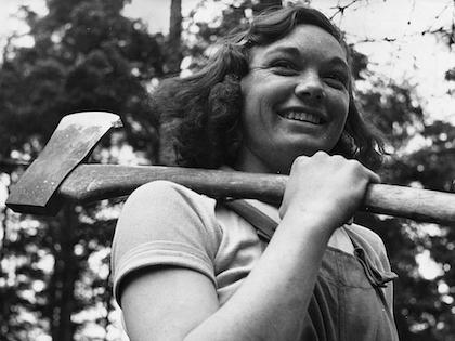 A black and white photo of a woman in overalls with an axe slung over her shoulder, smiling.