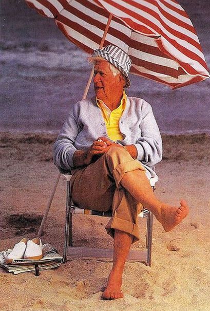 A picture of a barefoot Tip O'Neill on the beach in khaki pants rolled up above the thighs, a sweater with a yellow polo underneath, sunbathing under an american flag styled umbrella.