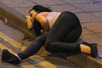 A picture of a woman laying on a curb using a slice of pizza as a pillow.